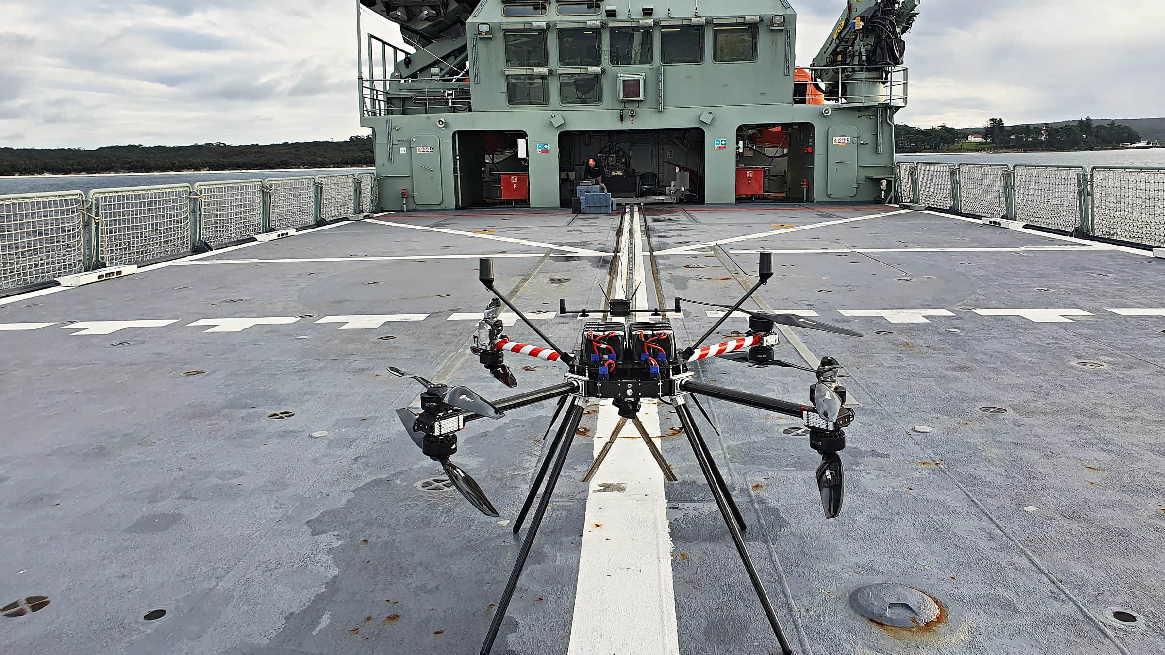A Callisto on the flight deck of a naval vessel, its arms extended, the ship's superstructure behind
