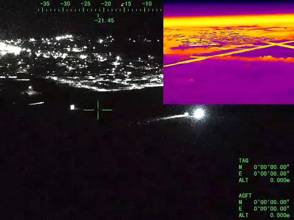 A night feed from the aircraft's gimbal camera tracking a vehicle's lights on a road below a lit town, with a thermal view inset at the upper right