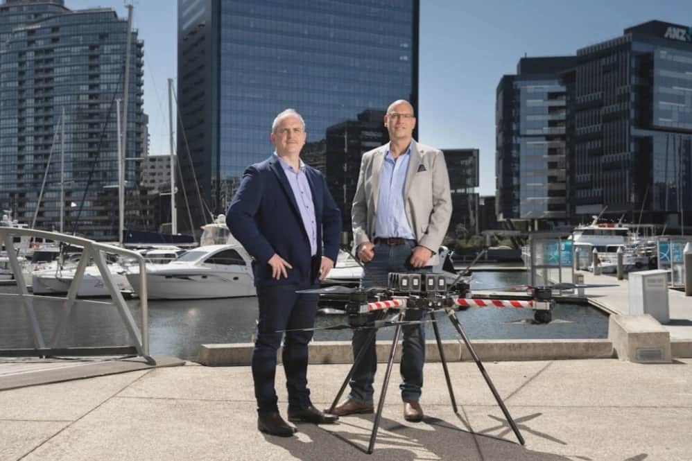Two men in suits standing either side of a Callisto on a waterfront promenade, office towers behind