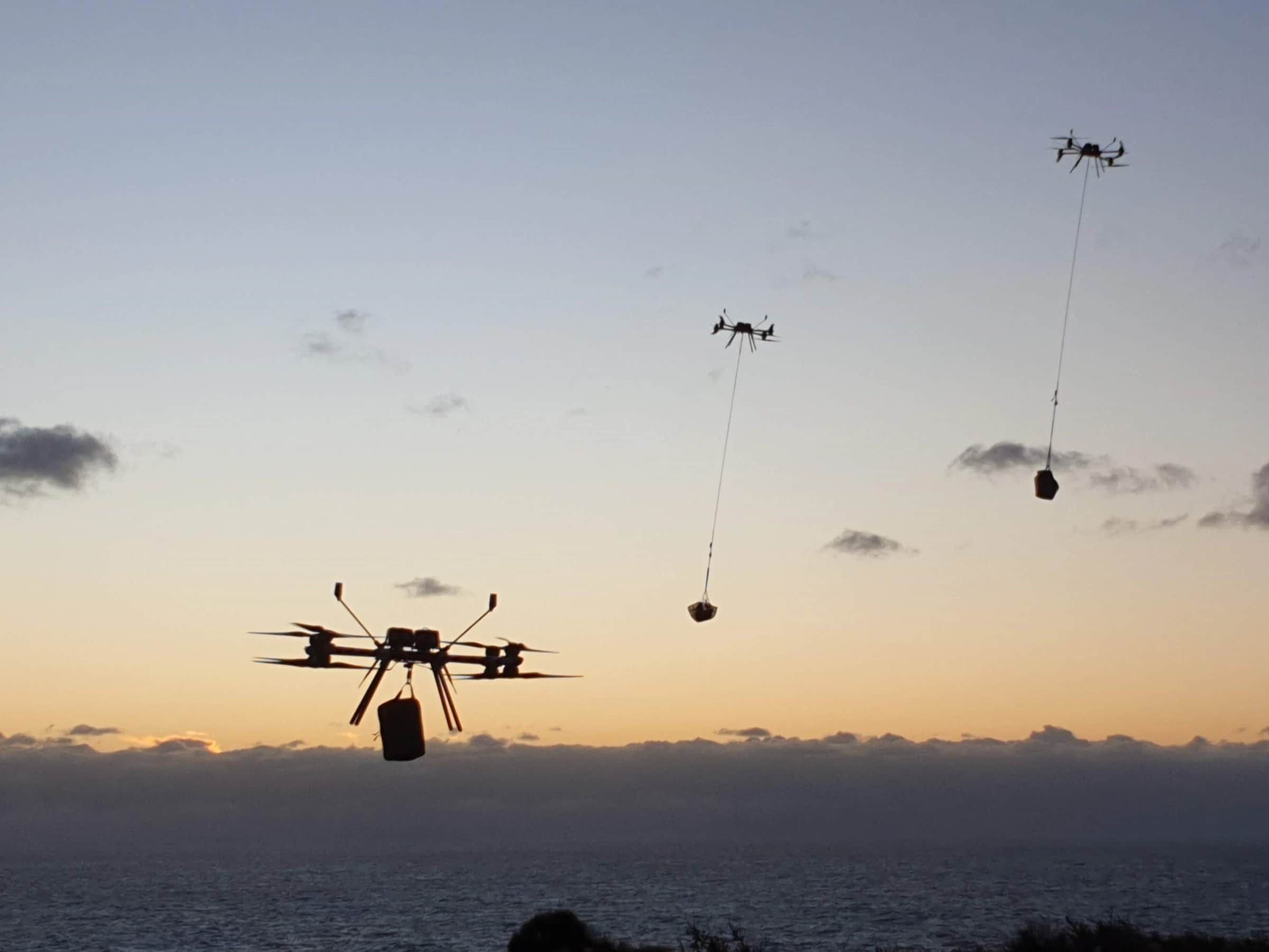 Three Callisto aircraft at dusk over the sea, each carrying a slung load