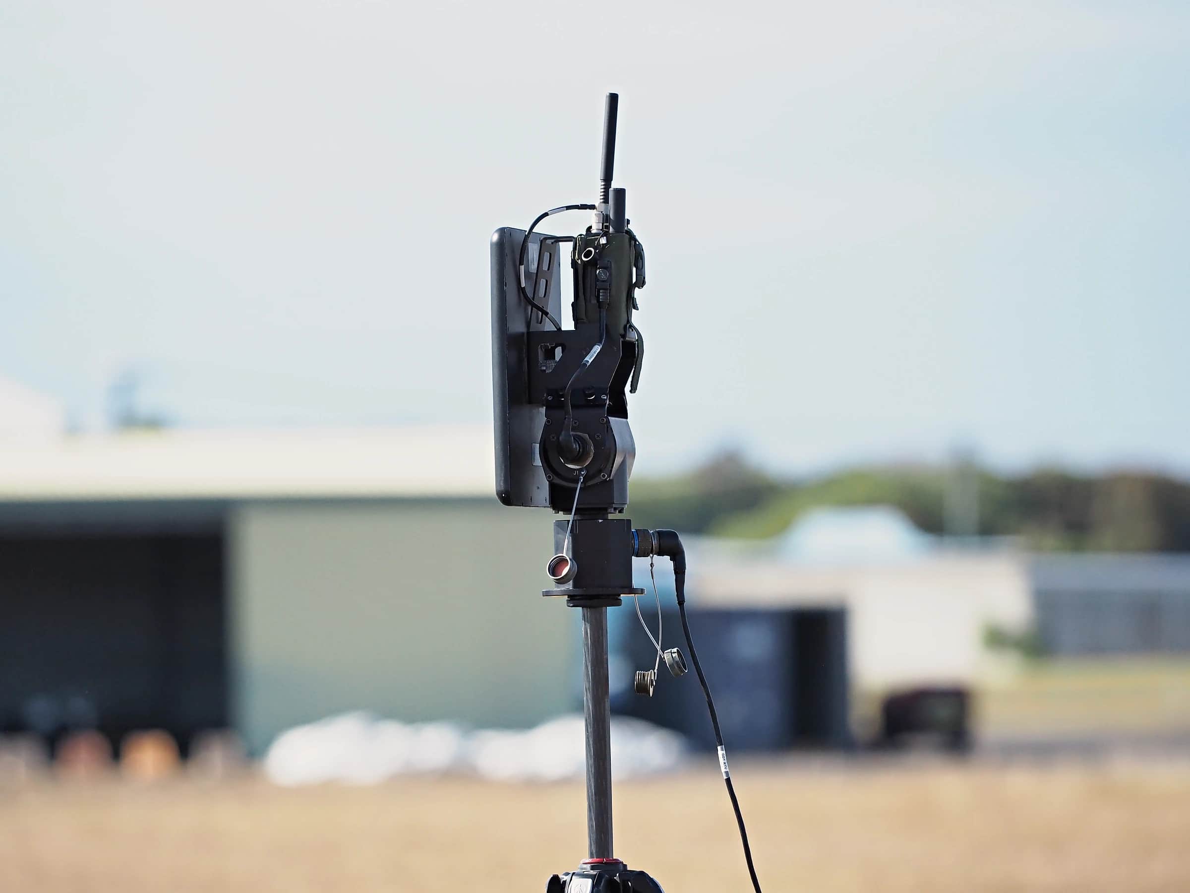 A communications relay mast on a tripod at an airfield, hangars behind