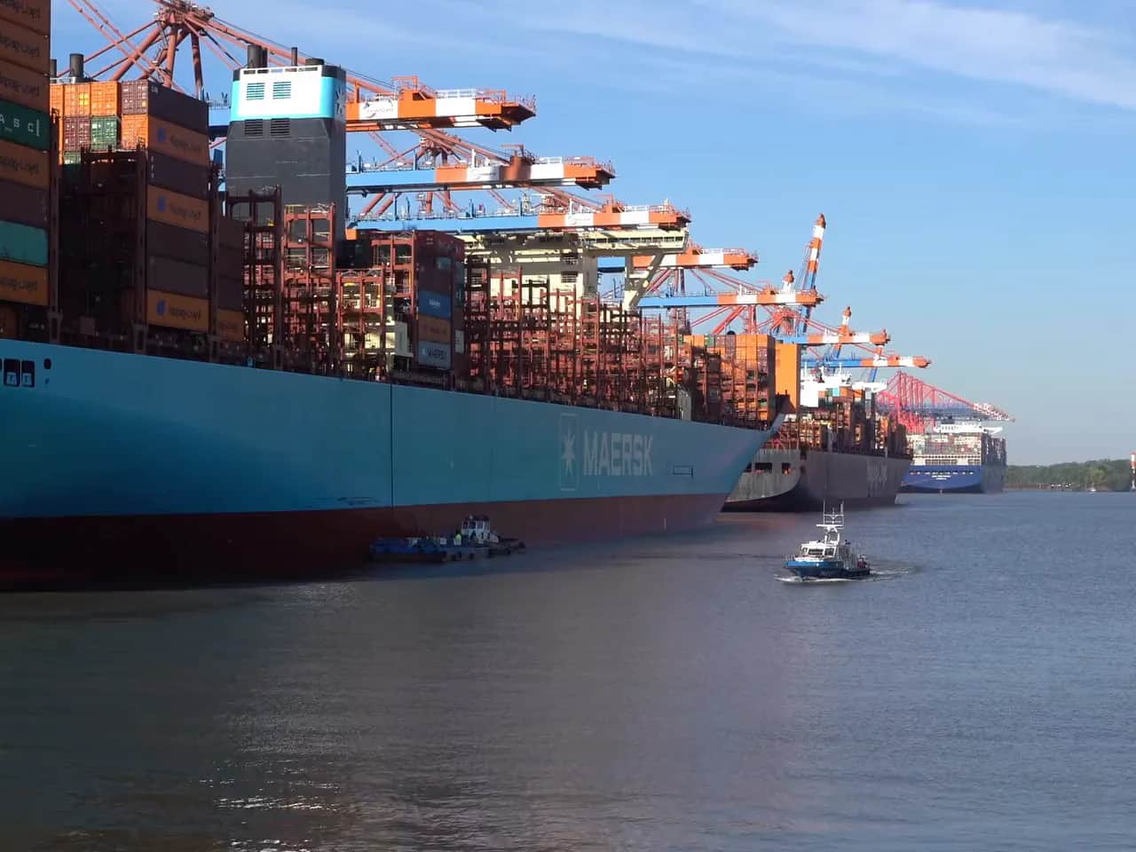 A tug working alongside a large container vessel at a container terminal, cranes stacked with containers above