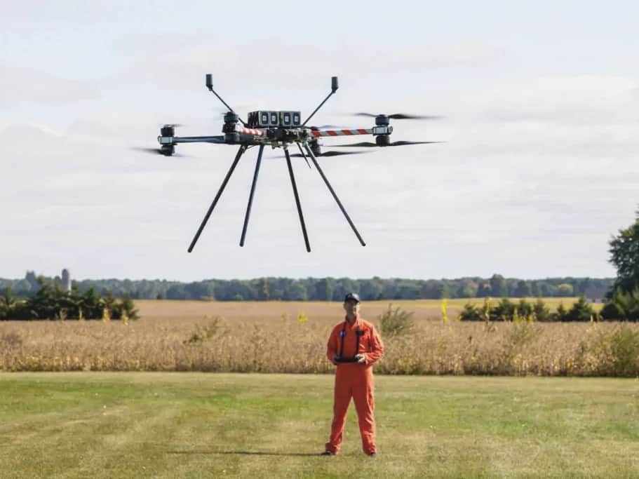 A pilot in an orange flight suit flying a Callisto over a grass field, farmland and trees behind