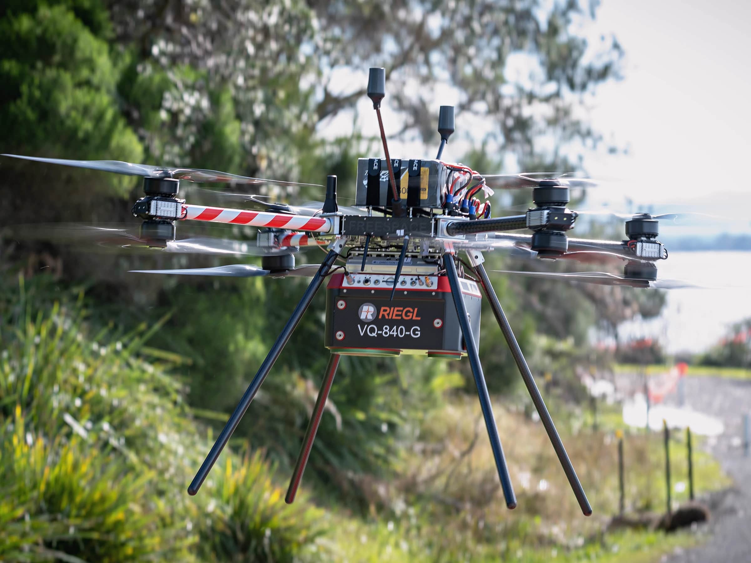 A Callisto in flight carrying the RIEGL VQ-840-G LiDAR, coastal scrub and water behind