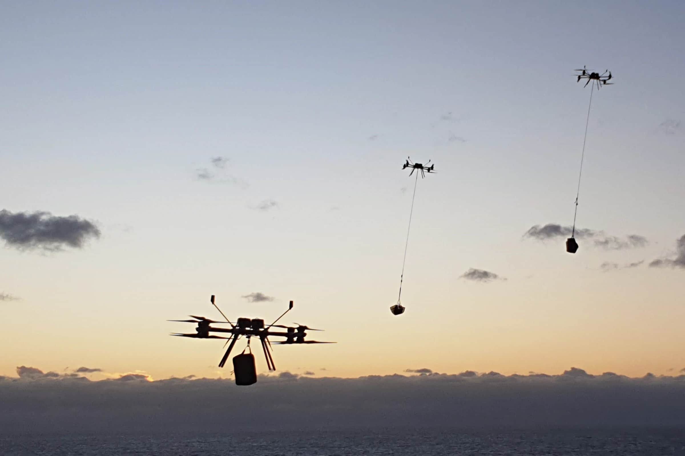 Two Callisto aircraft at dusk over the sea, each carrying a load on a line, a third aircraft in the distance